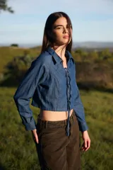 Camisa crop de denim azul con cuello camisero y cierre frontal mediante cordones ajustables a través de ojales. Presenta mangas largas con puños abotonados.