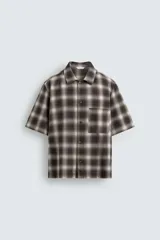 Sobrecamisa de corte relajado confeccionada en algodón, con estampado de cuadros en tonos marrones y beige. Presenta cuello solapa, manga corta, cierre frontal de botones y un bolsillo plastrón en el pecho.
