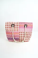 Bolso tipo tote con diseño de paneles en bloques que combinan estampado de lunares rojos sobre fondo blanco y franjas horizontales en tonos rosados, naranjas y morados. Cuenta con asas de soga gruesa de algodón en color crudo, sujetas con ojales metálicos y refuerzos de cuerina marrón.