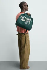 Bolso tipo shopper de algodón en color verde oscuro, con diseño de estampado tipográfico en color crema en el frente. Cuenta con dos asas de mano y cierre magnético.