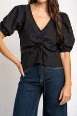 Blusa negra de algodón con diseño bordado sutil, escote en V y cordón ajustable fruncido en el centro del frente. Mangas cortas abullonadas.