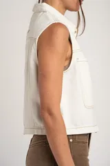Chaleco de denim blanco con diseño sin mangas, cuello camisero, bolsillos frontales con solapa y cierre de botones metálicos con detalle de ribete ondulado en contraste.