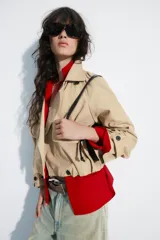 Trench corto de cuello solapa y escote en pico, color beige, con mangas largas terminadas en trabillas con botones. Cuenta con bolsillos laterales con solapa, cierre frontal cruzado con botón y bajo abullonado con elástico interior.