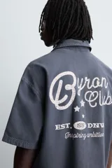 Camisa de algodón con estructura canvas, color rojo lavado, de corte relajado (relaxed fit) y manga corta. Posee cuello solapa, dos bolsillos de plastrón en el pecho y cierre frontal de botonadura. Presenta bordado y estampado de texto a contraste en el delantero.