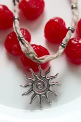 Collar de cordón de tela con estampado de cuadros vichy en tonos verde y rosa, con dije central de acero en forma de sol con acabado texturizado y detalles metálicos cilíndricos.