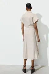 Vestido midi camisero de color beige, con cuello solapa, manga corta y cierre frontal con botones. Incluye un cinturón del mismo tejido con hebilla metálica.