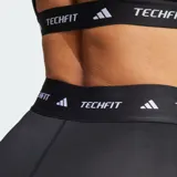 Calza capri deportiva Adidas Techfit de color negro, con diseño de compresión y tiro alto. Presenta una cintura elástica ancha con la inscripción "TECHFIT" repetida y el logo de la marca en blanco en la pierna izquierda.