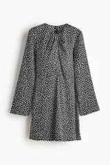 Vestido corto negro con estampado de lunares blancos, escote redondo con detalle girado y abertura frontal, mangas largas amplias con abertura en los puños, costura decorativa sobre la cadera y falda con leve vuelo.