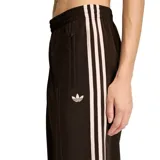Pantalón Adidas Firebird Loose de corte holgado y diseño deportivo, color marrón con las tres tiras características de la marca en los laterales y el logo del trifolio bordado en color rosado claro. Cuenta con cintura elástica y bolsillos laterales con cierre.