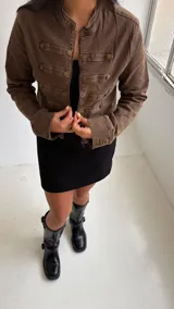 Chaqueta de estilo militar en color marrón, con corte estructurado y cuello mao. Presenta cierre frontal con cremallera y detalles de filas horizontales con botones metálicos decorativos en el frente y en los puños.