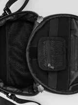 Bolso circular de estructura rígida, color negro, diseñado para el transporte de auriculares y accesorios. Cuenta con manija superior reforzada, correa de hombro ajustable con logo grabado, cierre perimetral y bolsillo trasero de acceso rápido.