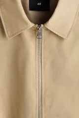 Campera tipo bomber de sarga de algodón en color beige, con cuello inglés, cierre frontal metálico y bolsillos diagonales ribeteados. Presenta terminaciones con elástico en los puños y el ruedo.