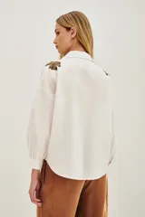Camisa blanca de algodón con cuello camisero y mangas largas abullonadas con puños ajustados. Presenta detalles bordados en los hombros con motivos vegetales y aplicaciones brillantes.