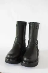 Bota de caña media confeccionada en cuero negro, con suela gruesa track antideslizante. Presenta un diseño estilo biker con estribo integrado, hebillas metálicas decorativas y una correa ajustable en la parte superior de la caña. Incluye cierre lateral para facilitar el calce.