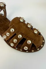 Sandalias planas de gamuza color tostado, con múltiples tiras decoradas con tachas metálicas doradas semiesféricas. Cuenta con una tira ajustable en el tobillo con hebilla metálica.