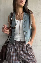 Chaleco de jean color celeste claro, con escote en V, sin mangas y decorado con tachas metálicas doradas distribuidas en los bordes y formando diseños en los paneles frontales.