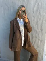 Blazer de gamuza color marrón, con solapas clásicas, cierre frontal de botones y bolsillos delanteros tipo parche.