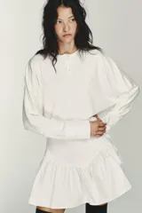 Vestido corto blanco con cuello redondo y manga larga tipo murciélago. El bajo tiene un acabado voluminoso con tejido combinado a tono y cierre frontal con botones.