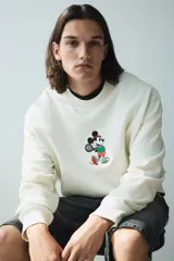 Buzo de tejido de algodón color crema, corte oversized y cuello redondo. Presenta un pequeño bordado central de Mickey Mouse vestido con ropa deportiva y sosteniendo una raqueta de tenis.