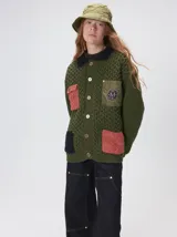 Campera tipo saco tejido de punto grueso en color verde oliva, con forro interno de nylon. Presenta cierre frontal con botones, cuello camisero en contraste y cuatro bolsillos delanteros con detalles en diferentes texturas y colores. Diseño holgado con aplique decorativo en la espalda.