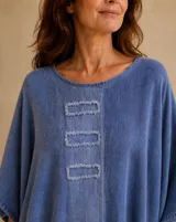 Blusa azul jean de corte holgado y asimétrico, con cuello redondo, mangas amplias y detalles de bordes deshilachados en el ruedo, mangas y en tres apliques rectangulares frontales.