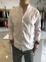 Camisa de manga larga color rosado, con cuello mao y cierre frontal de botones. Presenta un pequeño bordado dorado en el pecho y diseño de corte clásico.