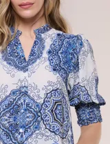 Blusa de crepe color blanco con estampado paisley en tonos azules y negros. Presenta cuello mao con escote en V, cierre frontal con botones y mangas cortas abullonadas con puños elastizados en punto smock.