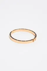 Pulsera rígida de metal dorado con detalles de esmalte negro en el borde y letras grabadas a lo largo de la pieza. Cuenta con cierre de bisagra.