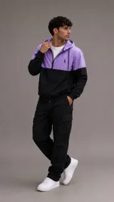 Conjunto deportivo compuesto por una campera rompevientos con capucha y cierre frontal, de diseño bicolor en violeta y negro, y un pantalón cargo negro con bolsillos laterales. La campera presenta puños elásticos y un pequeño logo bordado en el pecho.