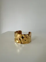 Brazalete rígido de acero quirúrgico con acabado dorado y diseño de superficie irregular con efecto ondulado.