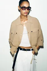 Chaqueta corta marrón de cuello solapa y manga remangada por debajo del codo. Presenta bolsillos delanteros de plastrón con solapa, bajo ajustable con cordón interior y cierre frontal con botones.
