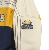 Buzo Adidas Varsity de diseño color block en tonos beige, amarillo y azul marino, con el logo clásico de la marca en el pecho y las clásicas tres tiras en las mangas.
