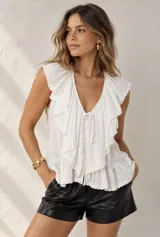 Blusa blanca sin mangas con escote en V, volados frontales amplios con bordado Inglés en los bordes, cierre frontal con botones y lazo ajustable.