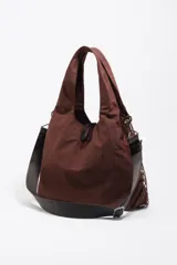 Bolso tipo tote de Bimba y Lola en color marrón, confeccionado en tejido técnico con un bolsillo frontal exterior cubierto por una red decorativa. Cuenta con asas superiores, correa ajustable extraíble y parche con logo de la marca en la parte inferior.