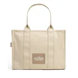 Bolso tote de lona en color beige, con diseño rectangular y estructura rígida. Presenta el nombre del modelo y la marca estampados en la parte frontal en color blanco. Cuenta con doble asa superior, cierre de cremallera en la parte superior y compartimento principal amplio.