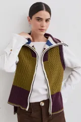 Chaleco de lana con cierre frontal completo y cuello alto. Presenta un diseño de bloques de color en tonos marrón chocolate y celeste, combinando secciones de tejido trenzado con secciones de tejido acanalado. Posee un corte asimétrico, siendo más largo en la parte posterior, y aberturas laterales en el ruedo.