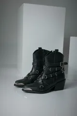 Bota corta texana de cuero color negro, con puntera metálica plateada, caña con cierre lateral y múltiples correas con hebillas y argollas metálicas.