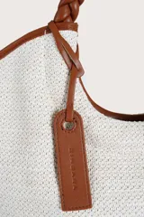 Bolso tipo tote de textura tejida en color blanco, con detalles de ribetes y asas trenzadas en color marrón. Incluye una etiqueta decorativa colgante en el mismo tono de las asas.
