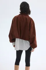 Campera corta marrón de tejido suave con terminación cepillada, cuello camisero, hombros caídos y cierre frontal. Cuenta con bolsillos laterales ribeteados y elástico revestido en puños y dobladillo.