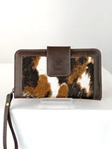 Billetera grande de cuero marrón con detalles en cuero texturado y panel central de pelo con estampado animal print tipo vaca en tonos blanco, marrón y negro. Cuenta con tira de muñeca y cierre de solapa con broche magnético grabado con el logo de un búho.