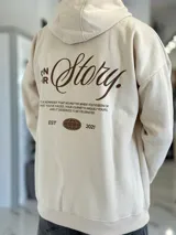 Canguro oversize de color beige con capucha y bolsillo tipo canguro al frente. En la espalda cuenta con un gran estampado tipográfico en marrón con la frase "Own Your Story", texto descriptivo y un logotipo ovalado con las letras "EST 2021".