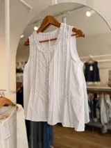 Blusa blanca sin mangas, confeccionada en tela liviana con tablitas frontales, cierre de botones al tono y cuello redondo decorado con un sutil volado fruncido.
