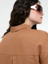 Camisa larga de lino color marrón, con corte oversize, mangas anchas y bolsillo frontal de cuero sintético en tono contrastante. Presenta cierre frontal con botones y diseño de bajo irregular.