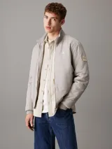 Campera Calvin Klein color gris claro, confeccionada en nailon 100% reciclado. Tiene cuello alto de canalé, puños y bajo elásticos, cierre de cremallera, bolsillos en la cintura, forro ligeramente acolchado y logo de la marca en el pecho y la manga.