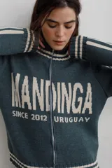 Campera tejida de algodón en color rojo, con cierre frontal completo, cuello alto y puños acanalados con detalles de rayas blancas. Presenta un diseño gráfico con texto blanco en el frente.