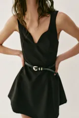 Vestido corto negro sin mangas, con escote en pico profundo y falda con pliegues. Incluye un cinturón fino de cuero sintético con hebilla metálica dorada en la cintura. Cierre lateral con cremallera oculta.