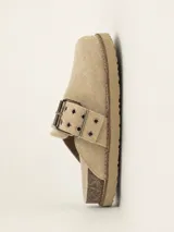Zueco de gamuza color beige, con diseño de punta cerrada y talón descubierto. Presenta una tira ajustable sobre el empeine con hebilla metálica y ojales decorativos. Cuenta con una suela de corcho y base de goma antideslizante.