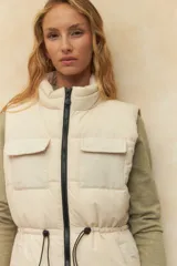 Chaleco puffer color crema, con cuello alto, cierre frontal y cuatro bolsillos con solapa. Cuenta con cintura ajustable con cordón.