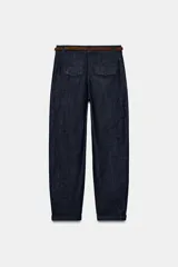 Pantalón chino de tiro bajo color marrón oscuro, con pernera recta y bajo con vuelta. Incluye un cinturón trenzado delgado de color marrón claro. Cuenta con bolsillos delanteros y cierre frontal con cremallera y botón.