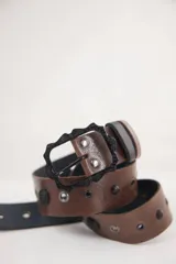 Cinturón de cuero marrón con hebilla rectangular de metal negro con diseño labrado. Presenta remaches metálicos negros de distintos tamaños a lo largo de la tira.